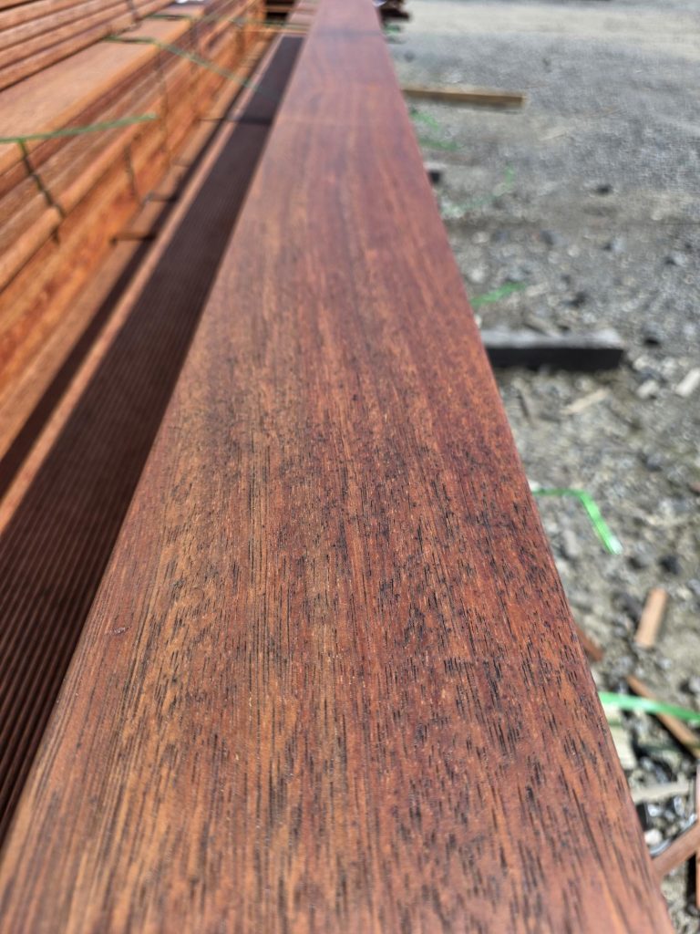 Close-up of reddish-brown wooden boards stacked outdoors, bound with straps on a gravel construction site